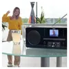 A woman is dancing in the background while a HAMA radio is on a table in the foreground. Produktbild-Vorschau