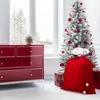 A red velvet Santa sack with white pompoms sits in front of a flocked Christmas tree decorated with red ornaments, next to a red dresser and a window showing a snowy landscape. Produktbild-Vorschau