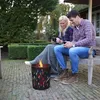 A couple sits on a bench next to a fire pit, holding mugs. Produktbild-Vorschau