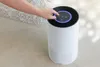 A hand is shown touching the control panel of a white air purifier. The air purifier is on a tiled floor next to a fluffy rug. Náhľad obrázku produktu