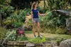 A woman is watering plants in a garden with a hose connected to a pump. Produktbild-Vorschau