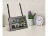A security camera monitor with four split screens is displayed on a white countertop, with a plant and a clock in the background. Produktbild-Vorschau