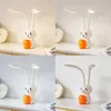 A collage of four images showing a white LED desk lamp shaped like a rabbit with an orange cup holder, displayed on a white desk with a clock in the background. The lighting and color temperature of the lamp vary across the four images. Produktbild-Vorschau