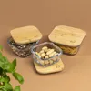 Three glass storage containers with bamboo lids are arranged on a brown background. One container is filled with nuts, another with pasta, and the third with what appears to be dried herbs or spices. A green leafy plant is visible in the lower left corner. Produktbild-Vorschau