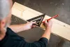A person is using a black triangular ruler and a red pencil to mark a wooden plank. Voir les images du produit