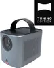 A portable projector is shown from a three-quarter front view against a white background. A black handle is attached to the top. A black square logo with white text is in the upper right corner. Podgląd zdjęcia produktu