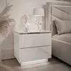 A white, glossy nightstand with two drawers next to a bed. Produktbild-Vorschau