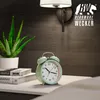 A mint green analog alarm clock sits on a white nightstand next to a potted aloe vera plant and some books. A wall lamp is visible in the background. Produktbild-Vorschau