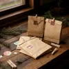 Brown paper bags with Christmas-themed stickers and wooden clothespins are arranged on a wooden surface, with pine branches in the background. Produktbild-Vorschau