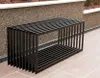 A modern black metal garden bench is placed outdoors on a paved patio, with a textured stone wall and landscaping in the background. Anteprima dell’immagine prodotto