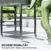 A person is pushing a black grill cart with three shelves on a paved patio with plants in the background. Produktbild-Vorschau