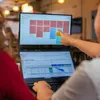 Two people are using a dual-screen laptop setup in a modern office or cafe environment. One person is pointing at the top screen, which displays a colorful grid of sticky notes, possibly a project management board. The bottom screen shows a Gantt chart, in Voir les images du produit