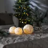 Three illuminated snowball-shaped candles are arranged on a textured tablecloth in front of a decorated Christmas tree. Produktbild-Vorschau