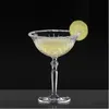 A crystal cocktail glass filled with a pale yellow drink, garnished with a lime slice and a salt rim, set against a dark background. Produktbild-Vorschau