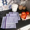 A kitchen counter with several folded tea towels, a garlic keeper, and sliced tomatoes on a cutting board. Produktbild-Vorschau
