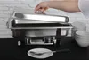 A hand is shown lifting the lid of a chafing dish. Several bowls are stacked to the right, and a plate with cutlery is in the foreground. Voir les images du produit
