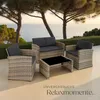 A rattan lounge set with dark cushions and a black glass coffee table is set up on a patio overlooking the ocean, with trees in the background. Náhled obrázku produktu