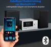A hand holding a smartphone displaying an audio app, with a compact audio system and speakers in the background, illustrating wireless music streaming via Bluetooth. Produktbild-Vorschau