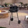 A charcoal grill is set up on a paved patio next to a fire pit, with outdoor furniture in the background. Produktbild-Vorschau