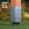 A metal post anchor is shown installed in the ground, supporting a wooden post. The background is a garden setting with grass and foliage. Produktbild-Vorschau