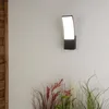 A modern black outdoor wall light with a curved white diffuser is mounted on a textured white wall. In the foreground, decorative grasses and plants add a natural element. Anteprima dell’immagine prodotto