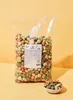 A large bag of mixed snacks with a small bowl of snacks in the foreground, set against a yellow background. Produktbild-Vorschau