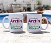 Two white ceramic mugs with German text and graphics are displayed on a wooden surface with a blurred background of a pool and plants. Produktbild-Vorschau