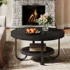 A round black coffee table with a lower shelf is placed in front of a stone fireplace with a fire burning. Oranges are on a plate on the table, and a vase of flowers is also present. A rug is under the table. Produktbild-Vorschau