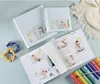 A baby album is open, showing photos of a baby, with baby toys and a blanket nearby. Voir les images du produit