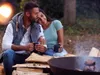 A couple sits by a campfire, holding mugs. Anteprima dell’immagine prodotto
