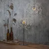 Two starburst-shaped LED lights are displayed against a textured gray wall. One light is on a stand in the foreground, and the other is suspended in the background. Dried reeds in brown bottles are to the left. Produktbild-Vorschau