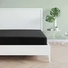 A black fitted sheet on a white bed frame, with a white headboard and a light green wall in the background. A small side table with plants and books is visible to the right. Produktbild-Vorschau
