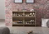 A shoe cabinet with multiple shelves and flip-down doors, filled with various shoes, is placed against a brick wall. A large canvas bag and a pouf are also visible in the scene. Produktbild-Vorschau