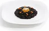 A close-up shot of a black rice dish with a raw egg yolk in the center, garnished with small pieces of orange carrot and herbs, served on a white plate. Anteprima dell’immagine prodotto