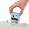 A hand is holding a blue stamp with a cartoon face over a white t-shirt, stamping a name and a rocket icon onto the shirt. Podgląd zdjęcia produktu