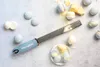 A Microplane grater is shown with macarons and pieces of cheese on a light gray surface. Náhled obrázku produktu