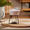 A pink wax burner with a white wax melt sits on a black metal stand, placed on a woven placemat with a lit tea light underneath. A potted fern is visible in the background. Produktbild-Vorschau