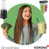 A young girl is holding a bunch of broccoli in one hand and a smoothie in a glass in the other, with a blender and two smoothies in to-go cups in the background. Produktbild-Vorschau