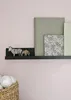 A black floating shelf mounted on a light pink wall, displaying decorative items including a zebra figurine, an elephant figurine, and two books. Produktbild-Vorschau
