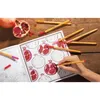 A hand is coloring a page in a book with pomegranates, surrounded by coloring pens and a real pomegranate. Produktbild-Vorschau