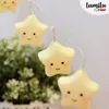 A string of yellow star-shaped LED lights with cute faces, hanging in front of a blurred green plant. Náhľad obrázku produktu