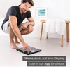 A man is crouching on the floor next to a body analysis scale, pointing at its display. Produktbild-Vorschau