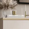 A close-up of decorative items on a shelf, including a vase with dried flowers, books, a candle, and candles in a holder. Náhled obrázku produktu