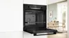 An open black built-in oven is shown in a modern kitchen setting. The oven door is pulled down, revealing the interior with racks and a fan. The background shows a dining area with a table and chairs. Produktbild-Vorschau