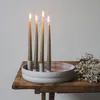 A round, light gray candle holder with four taper candles is placed on a rustic wooden table, with a small bunch of dried flowers to the right. Produktbild-Vorschau