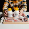A person is holding two bottles of varnish over a wooden board with a painted autumn scene. A paint roller and brush are also visible. Produktbild-Vorschau