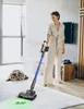 A woman is vacuuming a rug with a cordless vacuum cleaner, while a cat lies on the rug nearby. Produktbild-Vorschau