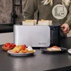 A person is using a toaster in a kitchen setting, with food items like waffles and tomatoes displayed on the counter. Voir les images du produit
