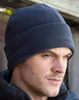 A man wearing a dark blue fleece hat and jacket, with a wooden structure in the background. Produktbild-Vorschau