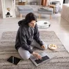 A woman is sitting on a rug in a room, using a laptop. A robot vacuum cleaner is visible in the background. Produktbild-Vorschau
